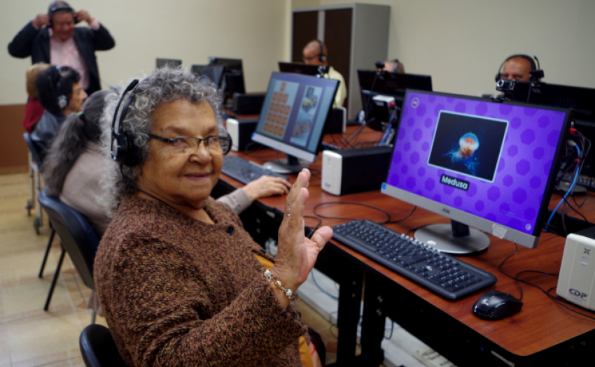 Adulta mayor en una sala de cómputo saluda hacia el frente. 