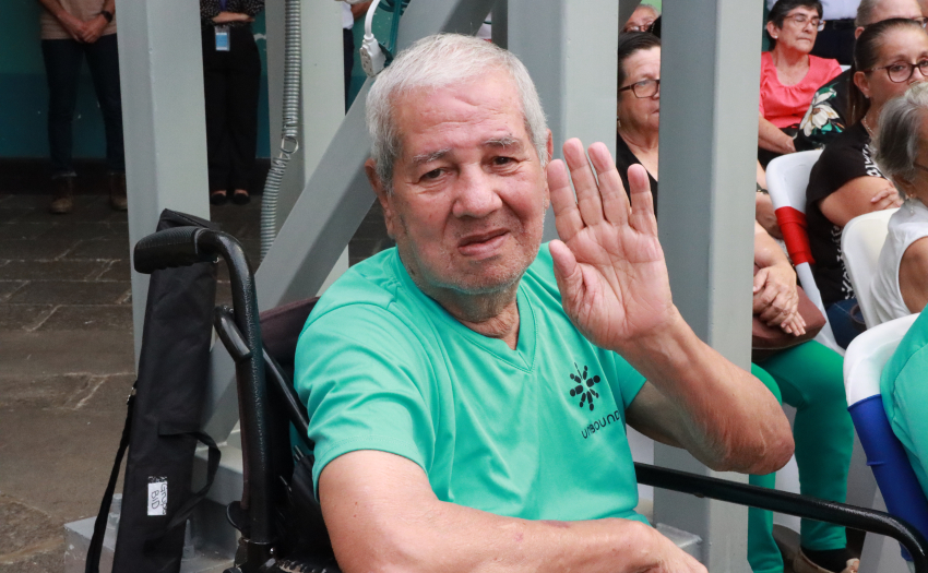 Adulto mayor en silla de ruedas saluda hacia el frente. 