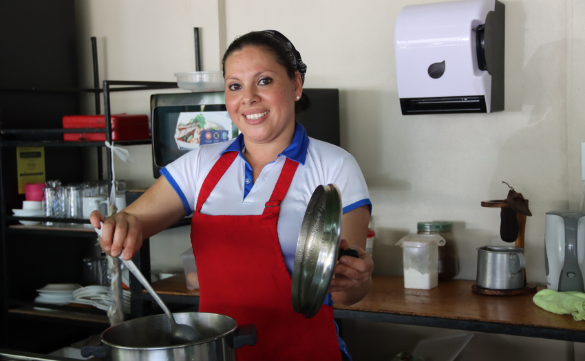 Emprendedora beneficiaria del IMAS. Está en su soda, coloca una cuchara en una olla. 